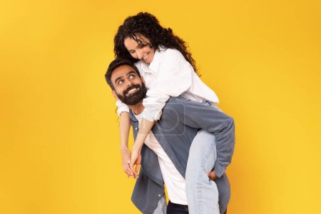Téléchargez les photos : Homme arabe joyeux portant sa petite amie sur le dos, Piggybackking femme aimante pour le plaisir, Poser et tromper ensemble sur fond jaune. Couple s'amuser, Studio Shot. Amour et bonheur - en image libre de droit