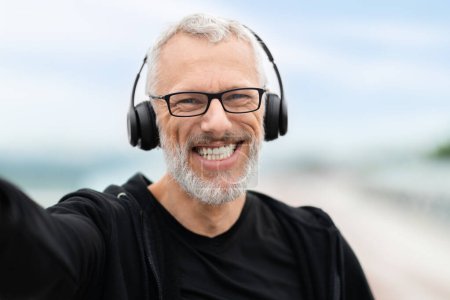 Cheerful handsome grey-haired elderly man wearing black sportswear fitness blogger taking selfie while jogging outdoors by footbridge in the morning, using wireless headphones, listening to music