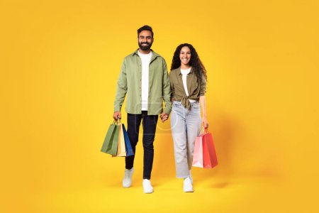 Téléchargez les photos : Shopping et consumérisme. Heureux jeune homme et femme arabique décontracté tenant des sacs à provisions colorés, marchant en tenant la main et souriant à la caméra sur fond jaune. Saison de vente, offre au détail - en image libre de droit