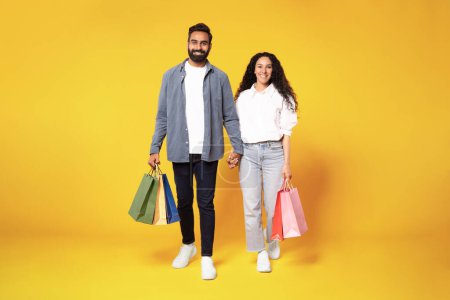Photo for Big Sales. Full Length Of Happy Arab Couple Walking With Shopping Bags In Studio On Yellow Background. Smiling Young Middle Eastern Man And Woman Holding Hands Carrying Purchases - Royalty Free Image