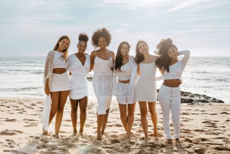 Foto de Hermosas señoras jóvenes multirraciales posando en la playa cerca del océano, teniendo noche de soltera, despedida de soltera. Grupo de mujeres abrazando y sonriendo a la cámara - Imagen libre de derechos