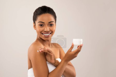 Photo for Attractive cheerful young black woman wrapped in white bath towel using body moisturizer after shower, holding white jar with cream, applying lotion on her shoulder, isolated on gray background - Royalty Free Image