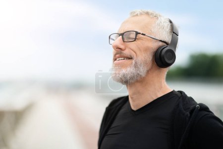 Téléchargez les photos : Entraînement extérieur. Joyeux homme âgé barbu aux cheveux gris en tenue de sport noire faisant de l'exercice à l'extérieur, faisant une pause, écoutant de la musique via un casque sans fil, profitant de l'air frais, copiant de l'espace - en image libre de droit