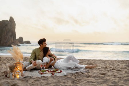 Téléchargez les photos : Pique-nique avec vue. Jeune couple romantique ayant rendez-vous sur la plage de sable près de l'océan, embrasser et profiter du temps ensemble au coucher du soleil sur la côte, espace de copie - en image libre de droit