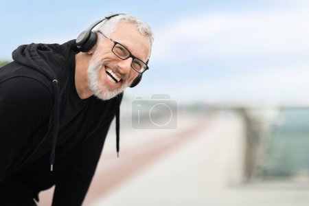 Foto de Primer plano de deportista de pelo gris sonriente positivo teniendo un descanso mientras trota, usando auriculares inalámbricos, escuchando música mientras se prepara para maratón, espacio para copiar - Imagen libre de derechos