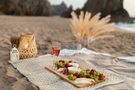 Foto de Picnic romántico con dos copas de vino de rosas o champán, frutas frescas y diferentes tipos de queso, fecha en la playa junto al océano al atardecer - Imagen libre de derechos