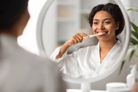 Foto de Hermosa mujer afroamericana joven cepillándose los dientes con cepillo de dientes cerca del espejo, mujer negra feliz sonriendo a su reflexión, disfrutando haciendo higiene matutina en casa, enfoque selectivo - Imagen libre de derechos