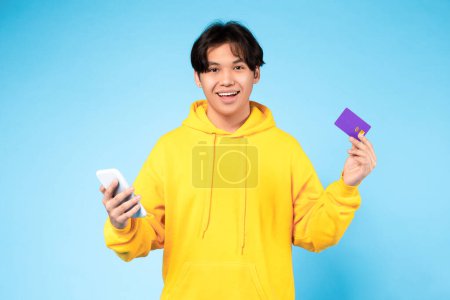 Photo for Online shopping app. Smiling young japanese teen guy using cellphone and credit card, purchasing in web store over blue studio background. Remote banking and internet sale offer concept - Royalty Free Image