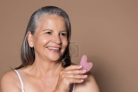 Photo for Cheerful elderly caucasian lady with gray hair shows jade stone for massage, enjoys anti-aging treatment, isolated on brown background, studio. Spa prosedure, beauty care in menopause - Royalty Free Image