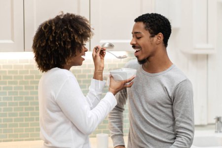 Photo for Loving young black woman with bushy hair feeding her handsome boyfriend, wife giving her husband oatmeal. Happy beautiful african american couple have breakfast together at kitchen - Royalty Free Image