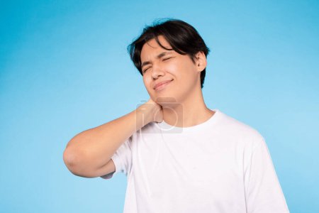 Photo for Portrait of tired asian student guy touching neck, feeling pain on blue studio wall background, posing with eyes closed. Unhappy boy massaging his neck. Sore muscles symptom and osteochondrosis - Royalty Free Image