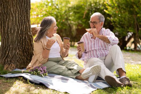 Téléchargez les photos : Heureux homme et femme âgés européens mangent des sandwichs, boire du café à emporter, profiter d'un rendez-vous romantique, déjeuner dans le parc, en plein air. Nourriture pour week-ends, vacances, amour et relations dans le temps libre - en image libre de droit