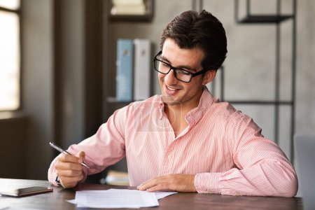 Photo for Handsome millennial european man in pink shirt sitting at desk at modern office. Young cheerful guy attending job interview, filling papers, survey and smiling, copy space - Royalty Free Image