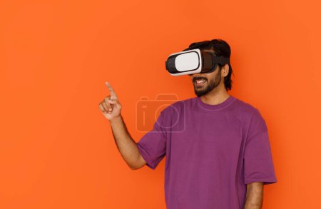 Foto de Hombre joven usando auriculares de realidad virtual, tocando el espacio de copia. Aislado sobre retrato de estudio de fondo naranja. VR, futuro, gadgets, tecnología, educación en línea, estudio, concepto de videojuegos - Imagen libre de derechos