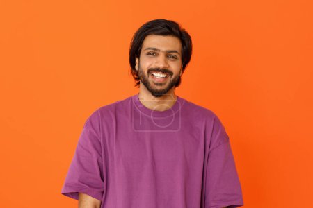 Photo for Happy cheerful positive handsome millennial eastern guy wearing casual purple t-shirt posing on orange studio background, smiling at camera, showing white teeth, copy space - Royalty Free Image