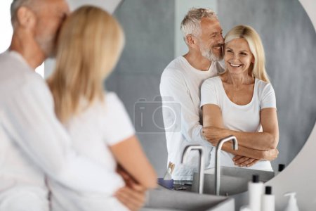 Téléchargez les photos : Heureux beau couple de personnes âgées regardant miroir dans la salle de bain et embrassant, Romantique Mature conjoints câlins tout en se préparant ensemble à la maison le matin, Focus sélectif sur la réflexion - en image libre de droit