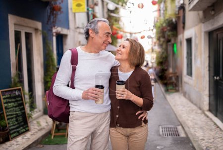 Téléchargez les photos : Aînés en vacances. Couple d'âge mûr romantique bénéficiant d'une promenade avec café, étreignant et souriant à l'extérieur, bénéficiant de l'amour sur une belle rue de Lisbonne. Touristes profitant de vacances au Portugal - en image libre de droit
