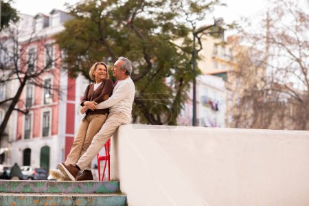 Photo for Romantic Senior Couple In Casual Outfits Sitting And Hugging In Urban Area Outdoor, Smiling To Each Other. Mature Man And Woman Enjoying Date Outside In City On Weekend. Copy Space - Royalty Free Image