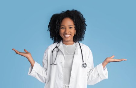 Cheerful young black lady doctor therapist in white coat hold free space in hands, isolated on blue studio background. Ad and offer, professional praxis, choice of health care