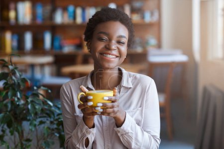 Photo for Out of home breakfast. Smiling beautiful African American millennial woman wearing smart casual outfit having coffee in cafe, holding yellow mug and smiling at camera, copy space - Royalty Free Image