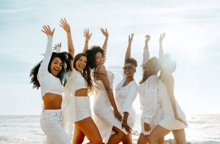 Foto de Grupo de amigas felices animando con los brazos levantados mientras celebran despedida de soltera en la playa, mirando y sonriendo a la cámara. Despreocupado equipo de novias mujeres jóvenes - Imagen libre de derechos