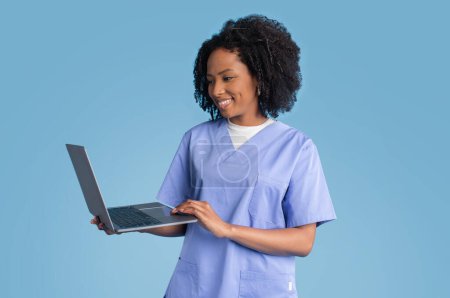 Téléchargez les photos : Heureux jeune infirmière médecin noir en uniforme bleu utilise ordinateur portable pour appel vidéo, isolé sur fond de studio bleu. Médecine, soins de santé, consultation à distance sur gadget en clinique - en image libre de droit