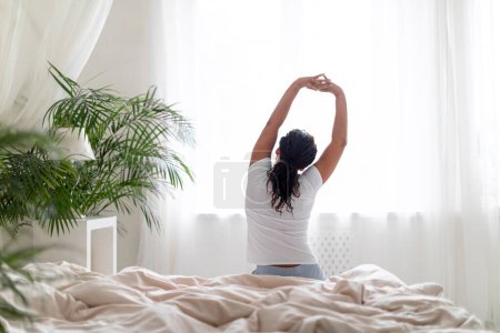 Photo for Young Black Woman Sitting In Bed And Stretching Arms After Waking Up, Rear View Of Female Relaxing At Home In The Morning, Resting In Bedroom After Good Sleep, Enjoying Lazy Day, Copy Space - Royalty Free Image
