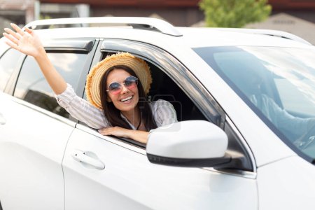 Téléchargez les photos : Jolie jeune femme excitée portant un chapeau en osier et des lunettes de soleil assises sur le siège avant, agitant la main par la fenêtre de la voiture et souriant à la caméra. Happy couple famille va vacances d'été en voiture ensemble - en image libre de droit