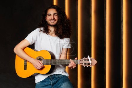 Foto de Happy Arab Musician Man Holding and Playing Acoustic Guitar Smiling At Camera At Black Wall In Studio (en inglés). Guy With Musical Instrument Plays His Songs (en inglés). Hobby y concepto de ocio - Imagen libre de derechos