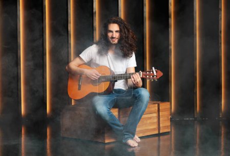 Photo for Professional Musician. Happy Hispanic Young Man With Curly Hair Playing Acoustic Guitar Sitting In Dark Studio With Smoke, Smiling Looking At Camera, Posing With Musical Instrument - Royalty Free Image
