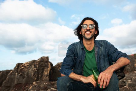 Téléchargez les photos : Beau jeune homme en lunettes de soleil se détendre en plein air sur des rochers de plage, sourire Millennial Guy tenant bouteille avec boisson gazeuse et regardant le ciel, profiter du temps ensoleillé et des loisirs à l'extérieur, Copier l'espace - en image libre de droit