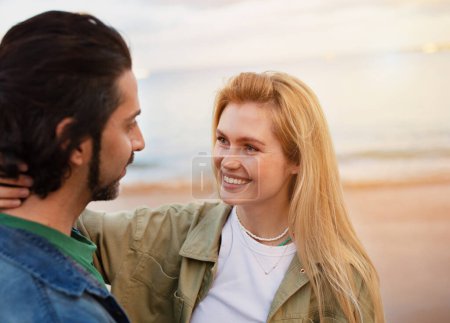 Foto de Feliz pareja joven abrazando mientras tiene una cita romántica en la playa, los amantes del milenio de pie en la orilla del mar, Disfrutando del ocio al aire libre, Mujer cariñosa mirando a su novio, Primer plano Shot - Imagen libre de derechos