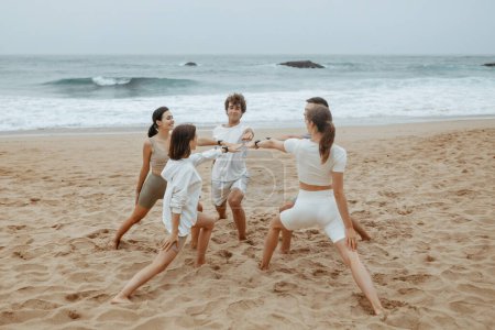 Téléchargez les photos : Brises de mer et poses guerrières. Groupe de jeunes en vêtements de sport faisant du yoga sur le bord de l'océan, debout en cercle, pratiquant des exercices d'asana sur la plage - en image libre de droit