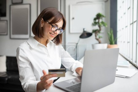 Photo for E-commerce offer. Happy businesswoman shopping online holding credit card and using laptop, sitting in coworking space. Internet banking payment service concept - Royalty Free Image