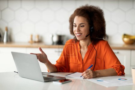 Photo for Online Lesson. Smiling Black Woman With Headset And Laptop Making Video Call At Home, Young African American Female Talking At Computer Webcamera And Taking Notes, Sitting At Table In Kitchen - Royalty Free Image