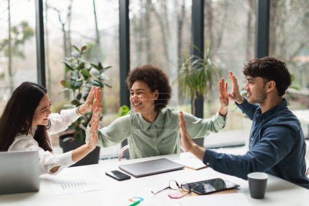 Photo for Celebrating of Unity. Diverse Young Business Team Shares High Five in Celebration of Shared Success And Achievement Sitting At Desk in Modern Office Interior. Triumph in Teamwork - Royalty Free Image