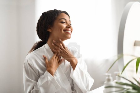 Photo for Neck Lift Concept. Smiling African American Woman In Silk Robe Touching Her Soft Smooth Skin While Looking In Mirror At Home, Happy African American Female Enjoying Result Of Beauty Treatments - Royalty Free Image