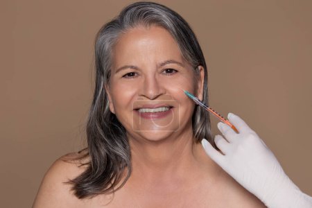 Photo for Glad old caucasian lady with gray hair enjoys anti-aging treatment uses injection from doctor, isolated on brown background, studio. Professional beauty care, wrinkle fighting - Royalty Free Image
