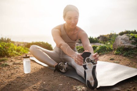 Foto de Joven atleta europea positiva en ropa deportiva haciendo ejercicio de estiramiento de piernas con botella de agua, disfrute del ejercicio en la playa de mar. Cuidado corporal, deporte y estilo de vida activo, fitness - Imagen libre de derechos