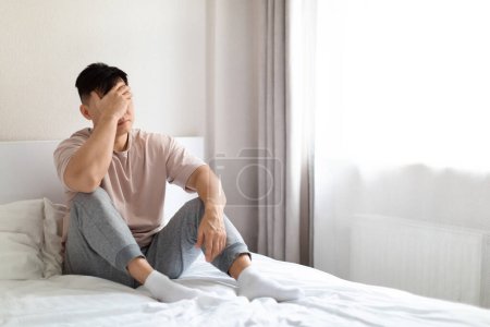 Photo for Frustrated unhappy depressed middle aged asian man wearing pajamas sitting alone on bed at home, covering his face, experiencing midlife crisis, going through divorce, copy space - Royalty Free Image