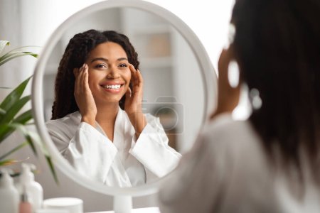 Téléchargez les photos : Concept de beauté. Portrait d'une jeune femme noire attrayante regardant le miroir et le visage touchant, belle femme afro-américaine bénéficiant de sa peau impeccable sans rides et souriant à la réflexion - en image libre de droit