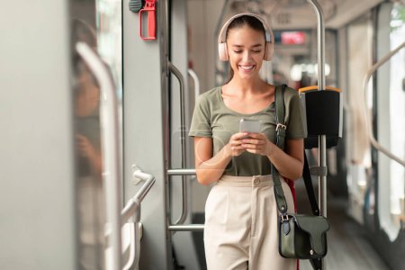 Téléchargez les photos : Transport en ville. Happy Passenger Woman Commuting debout dans le tramway et en utilisant un téléphone intelligent à l'intérieur, écouter de la musique en ligne via l'application et les écouteurs, profiter du transport en commun - en image libre de droit