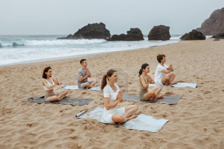 Téléchargez les photos : Formation de groupe. Hommes et femmes calmes excités méditant ensemble sur la plage au bord de l'océan, pratiquant le yoga en position lotus, assis sur des nattes et exerçant différents asanas, espace de copie - en image libre de droit