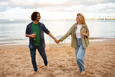 Téléchargez les photos : Aimer couple millénaire boire du vin tout en marchant sur la plage ensemble, heureux romantique jeune homme et femme tenant la main et se regardant, profiter de la date en plein air, pleine longueur prise de vue - en image libre de droit