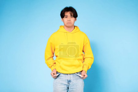 Photo for Poverty Concept. Upset Asian Young Teen Guy Showing His Empty Pockets And Looking At Camera, Having Financial Issues Standing Over Blue Studio Background. No Money, Crisis - Royalty Free Image