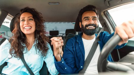 Téléchargez les photos : Joyeux couple moyen-oriental assis en voiture et tenant la main, profitant d'une promenade sur le siège passager avant et conducteur. Concept de propriété du véhicule - en image libre de droit