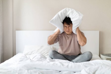 Photo for Unhappy irritated sleepless exhausted middle aged asian man wearing pajamas sitting on bed at home with closed eyes, covering head with pillow, suffering from strong migraine, copy space - Royalty Free Image