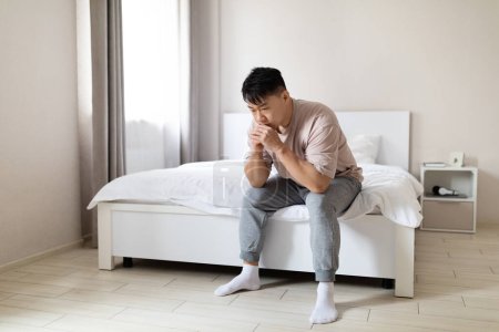 Photo for Frustrated unhappy depressed adult asian man wearing pajamas sitting alone on bed at home, leaning on his arms, have heavy thoughts, experiencing midlife crisis, going through divorce, copy space - Royalty Free Image