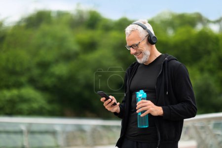 Photo for Senior sportsman drinking water, jogging outdoor. Elderly athletic man using smartphone and wireless headphones, choosing nice music for workout, holding bottle with water, copy space - Royalty Free Image