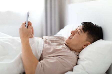 Photo for Happy handsome middle aged asian man wearing pajamas lying in bed at home, using cell phone and smiling, chatting with lover girlfriend, reading positive news, closeup - Royalty Free Image